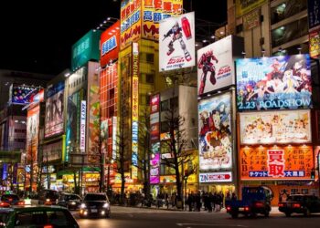 Vista noturna da vibrante Akihabara em Tóquio, iluminada por neons e anúncios de animes, com movimentadas ruas cheias de carros e pedestres