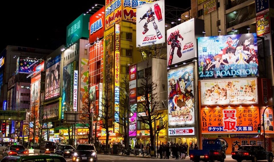 Vista noturna da vibrante Akihabara em Tóquio, iluminada por neons e anúncios de animes, com movimentadas ruas cheias de carros e pedestres