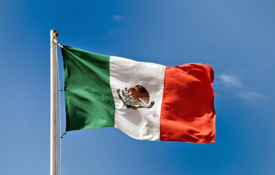 Bandera de México ondeando al viento bajo un cielo azul despejado, representando orgullo y patriotismo con el escudo nacional en su centro.