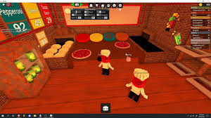 Roblox Work at a Pizza Place kitchen scene with players preparing pizzas on counters, surrounded by ingredients, ovens, and order boards in a brick-walled room.